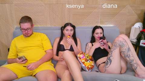 Video of three people on a couch: a man in yellow pajamas, a woman in a black bra, and a woman in a black tank top, eating snacks.