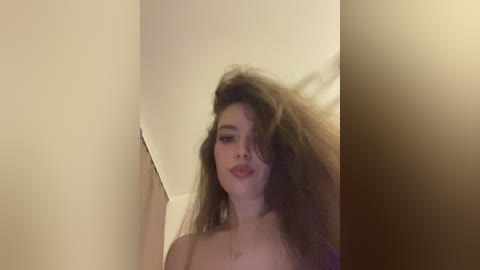 Video of a young Caucasian woman with fair skin and long, voluminous brown hair, partially covering her face. She wears a red lipstick and has a neutral expression. The background is a beige wall with a dimly lit, soft glow.