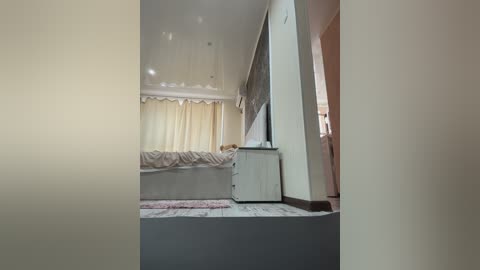 A video of a minimalist, modern bedroom with beige walls, a small bed, a white dresser, and a beige carpet. The room is well-lit with natural light filtering through sheer curtains.