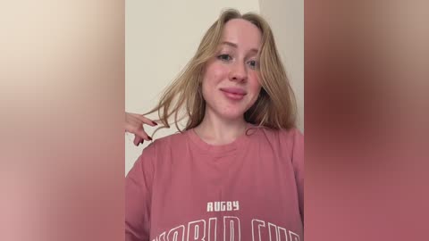 Video of a fair-skinned woman with long, wavy blonde hair, wearing a pink \"FUCK\" t-shirt, smiling and touching her hair.