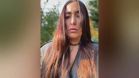 Video of a woman with long, straight brown hair, wearing a gray jacket, standing outdoors in a blurred green background.