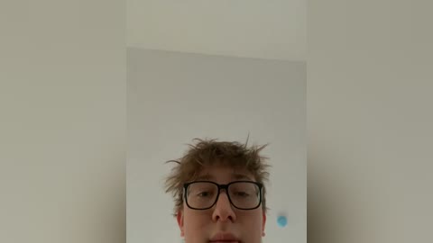 A video shows a young man with tousled hair, wearing black-rimmed glasses, standing against a plain white wall. He has a neutral expression.