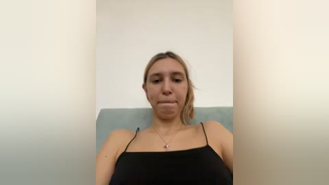 Video of a Caucasian woman with light skin and brown hair, wearing a black spaghetti-strap top, seated in a minimalist setting with white walls and a light blue cushioned surface.