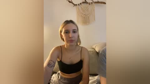 Video of a young woman with light skin and blonde hair, wearing a black spaghetti strap top and grey shorts, sitting on a grey couch, with a macram\u00e9 wall hanging and a tattoo visible on her arm.