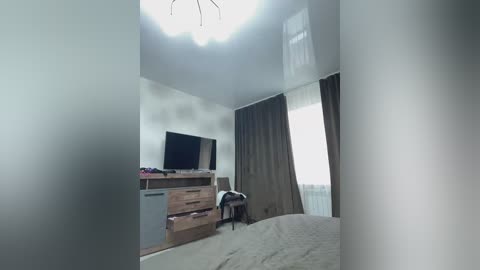 A video of a modern, minimalist bedroom with a large flat-screen TV on a wooden dresser against a white wall, a bed with light gray bedding, and sheer curtains covering a window.