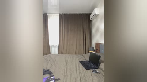 Video of a sparsely furnished bedroom with a beige bed, laptop on it, brown curtains, white wall, air conditioner, and a window with white blinds.