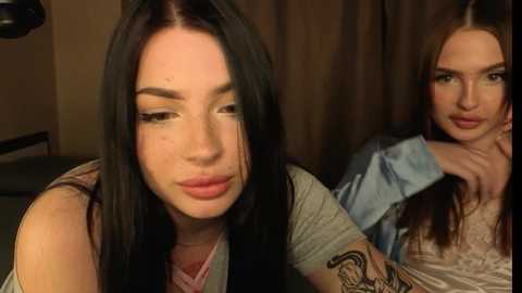 Video of two young women with long black hair, one with a tattooed arm and wearing a gray top, the other in a white top, indoors against a brown curtain background.