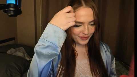 Video of a young Caucasian woman with long brown hair, fair skin, and freckles, wearing a light blue satin robe, adjusting her hair in a dimly lit bedroom with a gray bed and a brown curtain in the background.