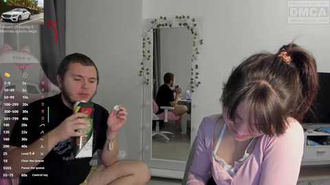 Video of a young man in a black T-shirt, sitting on a bed, holding a green bottle, and a young woman in a pink top, looking at a mirror, in a modern bedroom with a TV and a car in the background.
