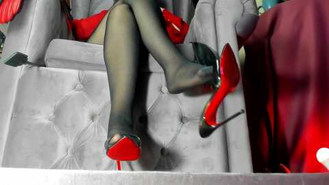 Video of a woman's legs in sheer black stockings and red-soled high heels, reclining on a gray, tufted sofa. Her long, red dress and a red handbag are visible, creating a sleek, luxurious atmosphere.