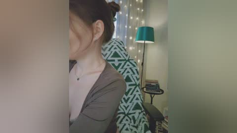 Video of a woman with fair skin and dark hair in a bun, wearing a grey cardigan over a white top, standing near a teal lamp and geometric-patterned curtains in a cozy room.