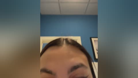 A close-up video of a woman with straight brown hair and light skin, seen from the top down, with blurred background featuring a blue wall and framed artwork.