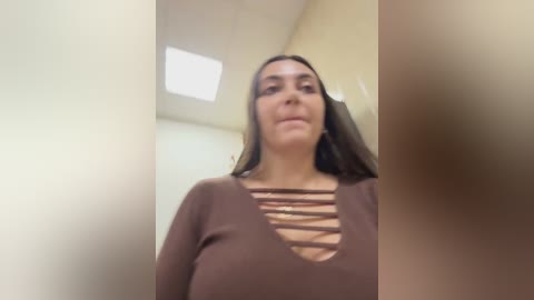 A video of a young woman with long brown hair, wearing a brown top with crisscross straps, standing in a dimly lit room with beige walls and a ceiling light.