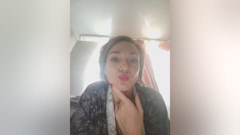 A video of a young woman with light skin, wearing a dark floral robe, making a kissy face in a small, dimly lit airplane cabin.