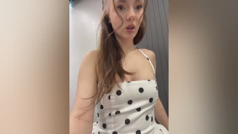 Video of a young woman with long brown hair, fair skin, and a slender build, wearing a white dress with black polka dots. She stands against a beige and grey background, looking slightly off-center.
