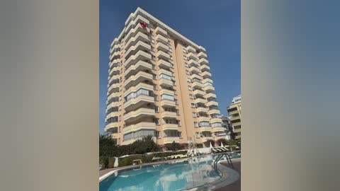 Video of a beige, modern, multi-story hotel with balconies under a clear blue sky. The pool in the foreground is surrounded by greenery and lounge chairs.