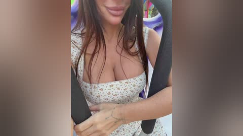 A video of a woman with long, dark hair and light skin, wearing a low-cut floral dress, sitting in a car with a black seatbelt across her chest.