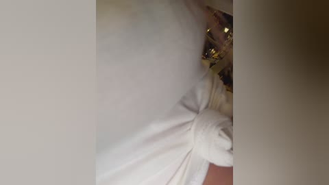 Video of a close-up of a person's arm in a white dress with a gathered waist, partially obscured by a blurred, gold-toned object, possibly a curtain or metallic fabric.