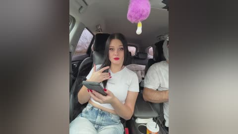Video of a young woman with long black hair, red lipstick, and white crop top, seated in a car with a purple duster hanging from the ceiling, holding a phone, against a gray interior.