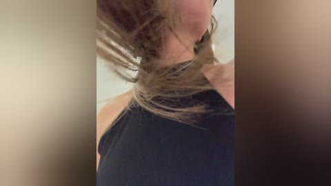 A close-up video of a woman with shoulder-length, messy brown hair, wearing a black sleeveless top, captured from the side, with a blurred, neutral-colored background.