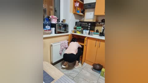 Video of a small, cluttered kitchen with a person in a pink shirt and black pants crouched beside a white refrigerator. Cabinets, microwave, and a small table are visible.