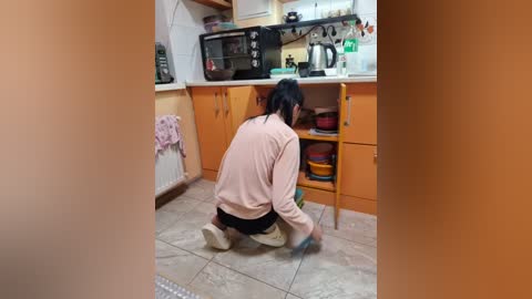 Video of a woman with black hair, wearing a pink sweatshirt, kneeling on a tiled floor, cleaning a small kitchen with orange cabinets, microwave, and a toaster.