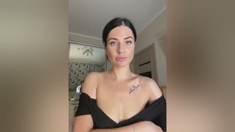 Video of a young woman with fair skin, dark hair, and a small tattoo on her shoulder, wearing a black off-shoulder top, standing in a modern bathroom with white walls and floral-patterned wallpaper.