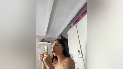 Video of a woman with medium skin tone and long dark hair, applying makeup in a small, brightly lit room with white walls and shelves.