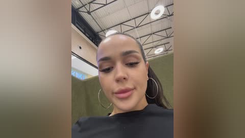 A video of a young woman with light brown skin and a slim figure, wearing a black top and large silver hoop earrings. She has long, straight hair tied back, and is indoors with a high ceiling and industrial lighting.