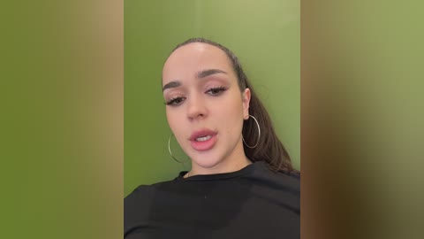 Video of a young woman with light brown skin, straight brown hair, wearing hoop earrings, and a black top, against a green background, with blurred edges.