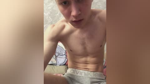 Video of a shirtless, lean young man with fair skin and short hair, wearing gray Calvin Klein boxer briefs, sitting on a bed with a floral-patterned sheet. The image is slightly blurred, focusing on the man's torso and face.
