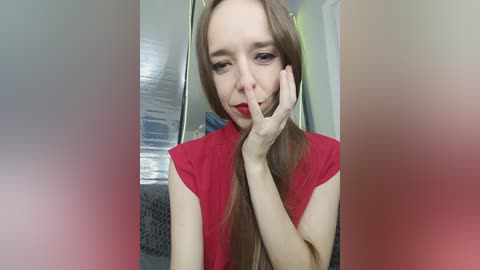 A video of a young woman with long, straight, light brown hair, fair skin, and wearing a red sleeveless top. She is covering her mouth with her right hand, smiling. Background shows a grey couch and a wall with a textured pattern.