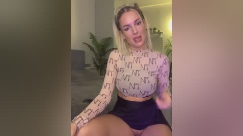 Video of a blonde woman with fair skin, wearing a long-sleeved crop top with \"NT\" pattern and black skirt, sitting indoors with a blurred background featuring plants and a couch.