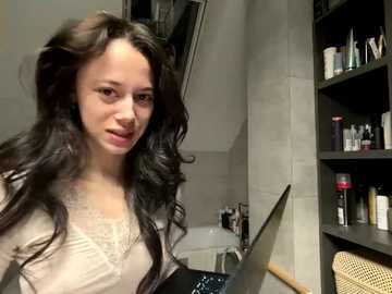 Video of a young woman with wavy, dark hair, wearing a beige lace top, standing in a bathroom with a mirror, shelves of toiletries, and a woven basket.