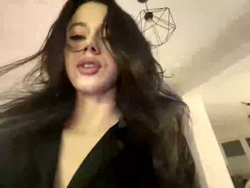 A video of a woman with long, dark hair, wearing a black dress, smiling, with a geometric pendant light in the background.