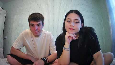 Video of a young man and woman sitting cross-legged on a bed. The man wears a white T-shirt, and the woman a black one. They both have long hair and are looking at each other.