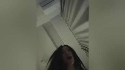 Video of a young woman with long dark hair, captured from a low angle, looking upwards. She has a distressed expression, standing in a dimly lit, narrow hallway with white walls and a gray ceiling.