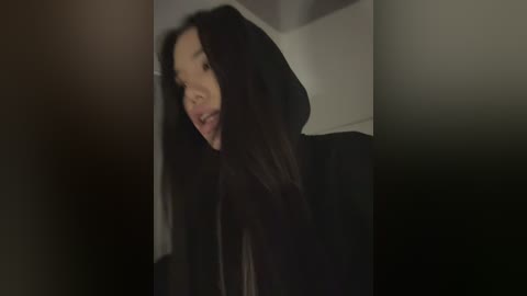 A video of a young Asian woman with long black hair and fair skin, wearing a black hoodie, stands in a dimly lit hallway. She has a contemplative expression.