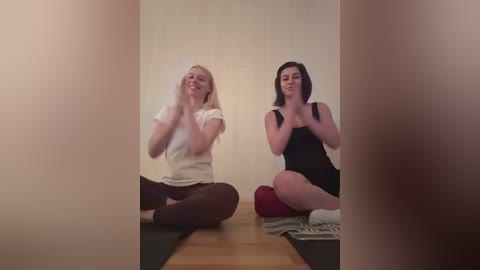 Video of two women meditating in a minimalist room, seated on a wooden floor with cushions. One wears a white top and brown leggings, the other a black tank top and black leggings. Soft, warm lighting.