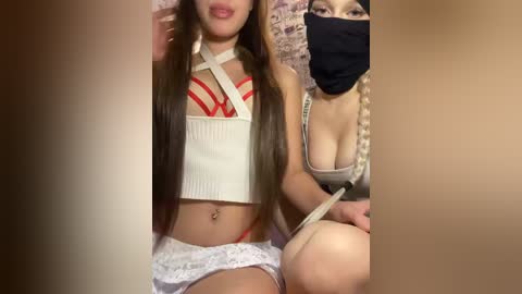 Video of two Asian women, one wearing a white ribbed crop top and red bralette, and the other a black mask and beige tank top.