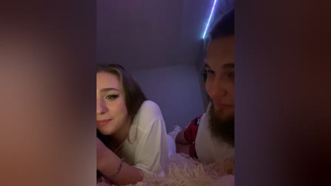 Video of two young women, one with light brown hair and one with dark hair, lying on a bed with a soft, textured blanket, smiling in dimly lit room with purple LED strip lighting.