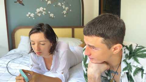 Video of a Caucasian man and woman in bed, he's shirtless with a necklace, she's in a white blouse, both holding smartphones, in a modern bedroom with a green wall and floral decor.