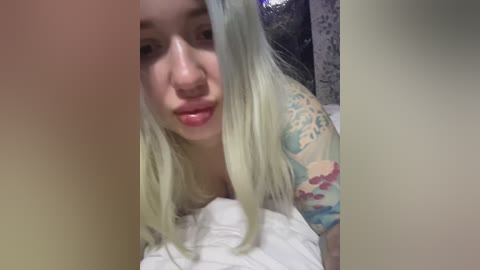 Video of a young, light-skinned woman with long, platinum blonde hair and a floral tattoo on her left shoulder. She has a neutral expression and is partially obscured by the frame.