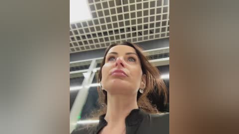 A video of a fair-skinned woman with long brown hair, wearing a black jacket, gazing upward with a thoughtful expression, against a modern, grid-patterned ceiling.