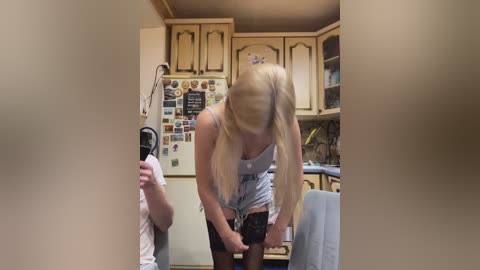 Video of a blonde woman in a tight, light blue dress and black stockings, adjusting her stockings in a cluttered kitchen with wooden cabinets, posters, and a blue chair.