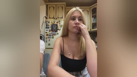 Video of a blonde woman with fair skin, wearing a black tank top and floral skirt, pensively touching her chin in a cozy kitchen with wooden cabinets, colorful magnets, and a poster.