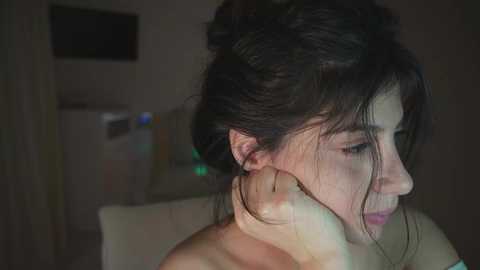 A dimly lit video of a woman with dark hair in an updo, leaning against a wall, with a man's hand on her shoulder. The background shows a blurred, dark room with a TV and a white couch.