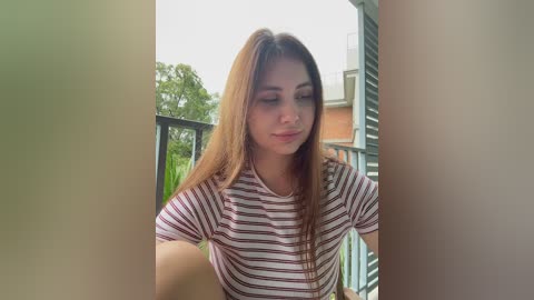 Video of a young woman with long brown hair, wearing a white and brown striped shirt, standing on a balcony with green foliage and a building in the background.