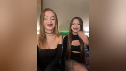 Video of two smiling young women with long hair, wearing black off-shoulder tops and chokers, in a dimly lit room.