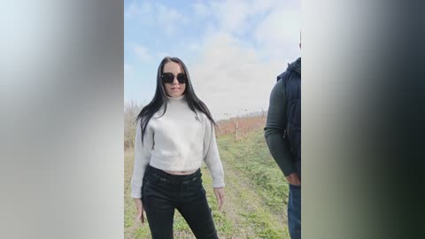Video of a young woman with long black hair, wearing a white turtleneck and black jeans, standing outdoors against a cloudy sky with green fields in the background.